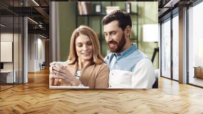 Portrait of young Caucasian joyful couple shopping on internet browsing online on tablet device sitting on sofa in apartment spending time together with cup of tea. Husband and wife resting at home Wall mural