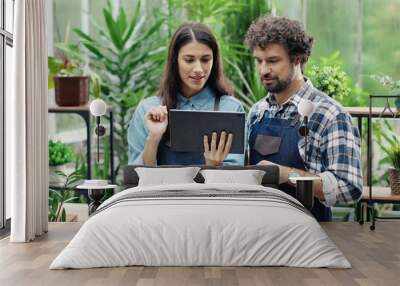 Portrait of young Caucasian lovely couple working in own flower shop together. Joyful female and male entrepreneurs in aprons typing on tablet and talking in floral store. Family business concept Wall mural