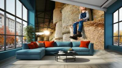 Pretty nice small Caucasian blonde girl smiling, resting and sitting on hay stocks in barn in August. Summer childhood in countryside. Little beautiful kid spending time in shed at farm. Happy child. Wall mural