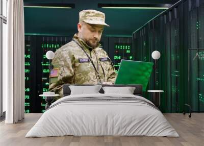 US army male Caucasian officer in uniform and cap stading at server wih secret data and using laptop computer to check the information. Military monitoring service room. Army networking concept. Wall mural