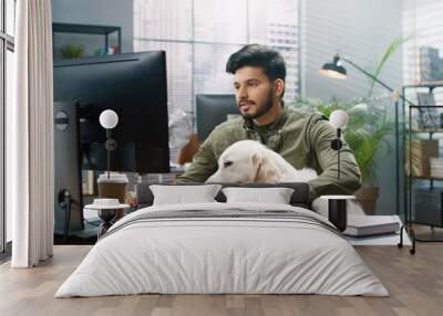 Young handsome Hindu guy sits in office with his big Labrador Retriever dog and works on computer completing task. Wall mural