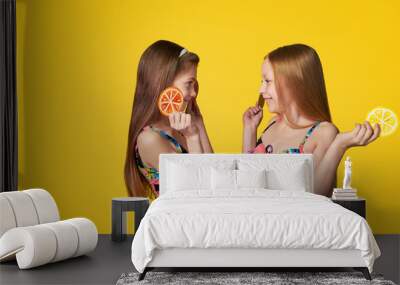 Portrait two cheerful girlfriends in colorful swimsuits looking at each other,holding image citrus.Caucasian teenager smile,enjoy vacation,summer season.yellow background.Fashion rest beach concept. Wall mural