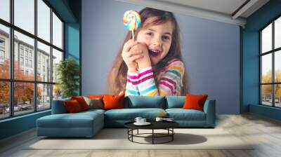 Little kid eating lollipop. Happy beautiful girl with candy isolated on blue background. Wall mural