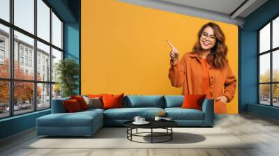 Woman in orange shirt pointing to the side with smile on her face. Generative AI. Wall mural