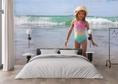 A beautiful little girl in a bright swimsuit and a hat stands against the backdrop of the sea on a bright sunny day, selective focus Wall mural