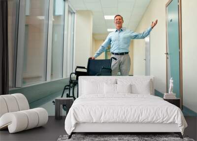 A handsome mature man stands recovered from his disability near a wheelchair and raised his hand in a sign of victory, standing in the clinic corridor. Wall mural