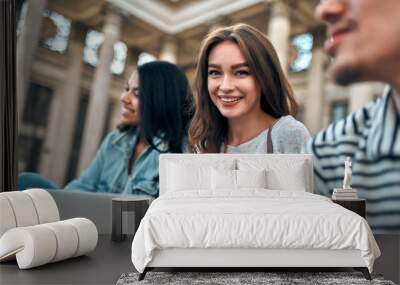 Attractive girl student with a laptop sits on the steps near the campus with her friends. Wall mural