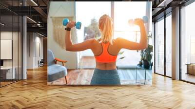 Determined woman losing weight at home and exercising with dumbbells. Sport and recreation concept. Beautiful woman in sportswear with blue dumbbells in her hands. Wall mural