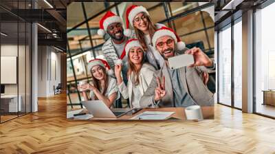 Happy New Year and Merry Christmas! Multiracial young creative people in Santa hats celebrate a holiday in a modern office and take a selfie with a smartphone on their last working day. Wall mural