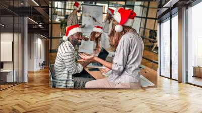 Happy New Year and Merry Christmas! Multiracial young creative people work in a modern office. A group of young business people are sitting in Santa hats on their last working day. Wall mural