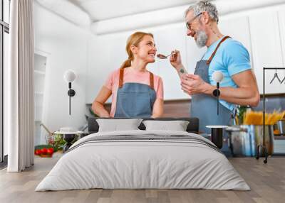 Happy senior couple in aprons are preparing pasta and fresh salad in the kitchen and having a nice time. Vegan, vegetarian, healthy lifestyle concept. Wall mural