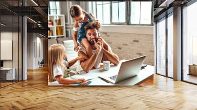 The businessman dad tries to work at the laptop when his little daughters play, fool around and interfere with him. Freelance, work from home. Wall mural