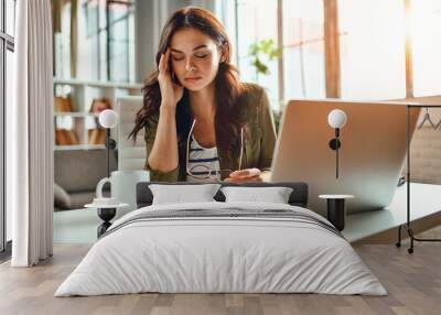 tired business woman in stress works at a laptop while sitting at a table at home and holds her hand Wall mural