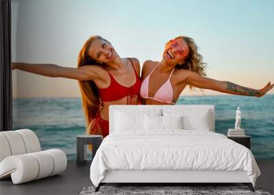 Two attractive women friends have fun on the beach in bikini swimwear at sunrise. Wall mural