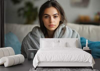 Serene young woman wrapped in a soft gray blanket, sitting on a modern sofa in a cozy living room Wall mural