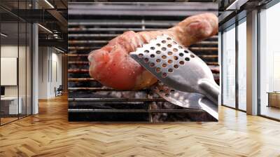 Metal tongues holding grilled sausage in front of bbq grill close up Wall mural