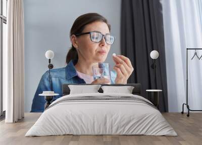 Mature middle-aged woman at home holding pill and glass of fresh water Wall mural