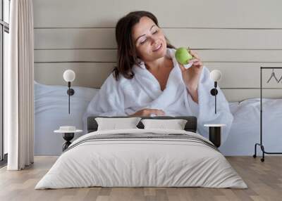 Middle-aged woman in white bathrobe with green apple in bed Wall mural