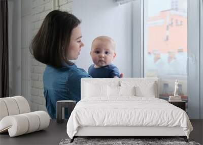 Mother holding baby in arms standing at home near window Wall mural