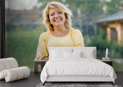 Outdoor portrait of positive confident mature woman. Smiling female blonde in a yellow dress with arms crossed near the house Wall mural