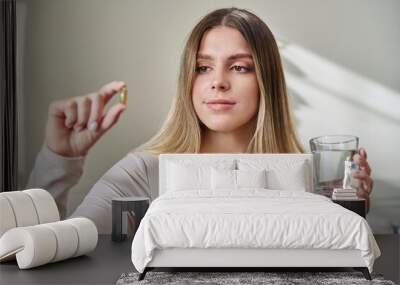 Young beautiful woman taking capsule with fish oil Omega 3 sitting on couch at home Wall mural