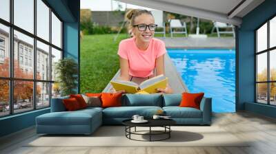 Young girl in glasses near the pool with a pile of books, reading book. Education, summer, knowledge Wall mural