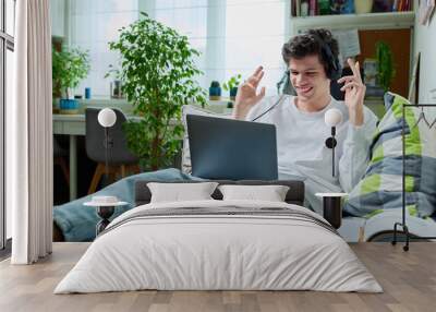 Young guy in headphones using laptop for video communication, lying at home Wall mural