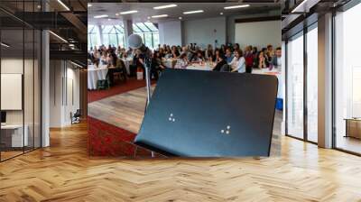 Atmosphere during corporate conference. A speaker's stand with microphone is viewed close up during a packed conference for professional people, blurry employees are seen sitting behind. Wall mural