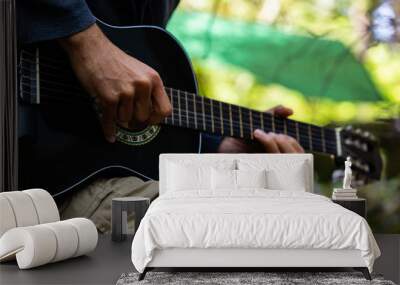 Close up and side view of a talented man, wearing shorts at a campsite during an earth festival, playing guitar with natural green bokeh in background Wall mural