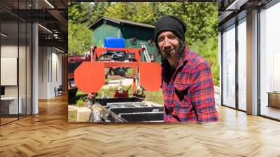 Close up portrait of a male lumberjack in mid thirties at work outdoors. Blurry machinery and cabin is seen in background with copy space to left. Wall mural