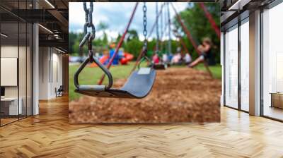 Closeup picture of a swing in a park for kids. Kids swigning in the blurry background - Picture taken on a warm summer day, with mulch ground instead of sand, and green lawn in the background Wall mural