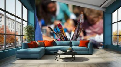 Kid is holding a metal container full of paintbrushes while sitting outside with other kids - Pictured at an artistic workshop in a family festival Wall mural