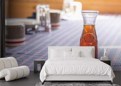 Maple syrup in glass bottle sitting on a table with a blue and red tablecloth, set for breakfast. Selective focus Wall mural