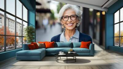 Smiling elderly woman, standing in the city. A happy old Caucasian grandmother wearing glasses, standing outdoors on a nice day. Good looking cheerful European senior female outside, close-up portrait Wall mural