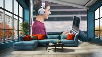 Smiling teenage boy with a laptop on the street. Handsome young man works on a notebook, outdoors. Cheerful guy holds a laptop on the knees and looking to screen. Teenager in headphones with laptop Wall mural