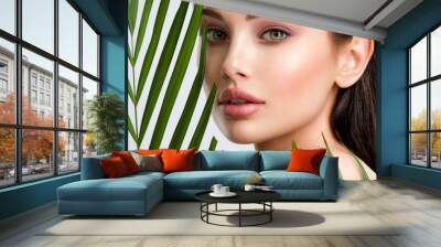 Young beautiful woman with healthy skin of face and palm leaves. Closeup fresh face of an attractive caucasian girl with green plants. Model with bright brown eye makeup. Skin care concept. Wall mural