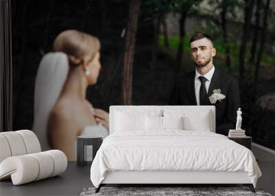 A bride and groom are standing on a balcony, with the groom looking at the bride. The bride is wearing a white dress and a veil, while the groom is wearing a black suit and tie Wall mural