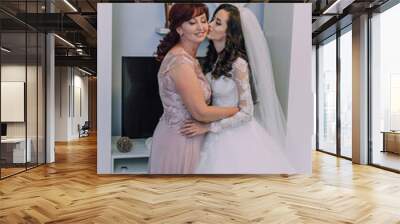 A bride and her mother are embracing each other in a living room. The bride is wearing a white wedding dress and a veil, while the mother is wearing a pink dress. The two women are smiling Wall mural