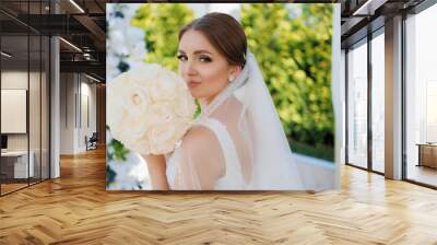 A bride is posing for a picture with a bouquet of white roses. She is wearing a white dress and a veil. Concept of elegance and romance, as the bride is dressed in a traditional wedding attire Wall mural