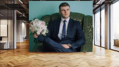 A man in a suit and tie sits in a green chair holding a bouquet of flowers. Concept of elegance and sophistication, as the man is dressed in formal attire Wall mural