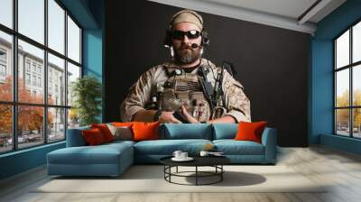 Brutal man in the military desert uniform and body armor stands and holds a gun on a black background in the Studio. The bearded player in the airsoft safety glasses or goggles and active headphones Wall mural