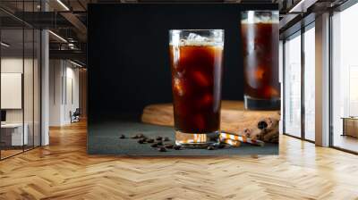 Ice coffee in a tall glass with cream poured over, ice cubes and beans on a old rustic wooden table. Cold summer drink with tubes on a black background with copy space Wall mural