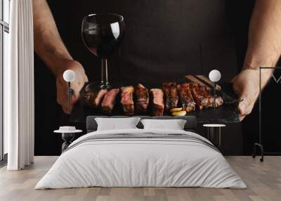 Man holding juicy grilled beef steak with spices and red wine glass on a stone cutting board on a black background Wall mural