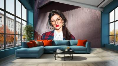 A brooding attractive girl with black hair, glasses and red lips, dressed in a plaid jacket and white shirt, stands near the wall in the sun and looks at the camera Wall mural