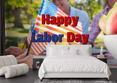 Composition of text happy labor day and face mask over happy family at picnic waving american flags Wall mural