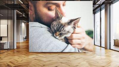 Close-up of cat and man. Portrait of a Devon Rex kitten and young beard guy. Handsome animal-lover man is hugging and cuddling his little kitty. Cat is purring and enjoying human company. Wall mural