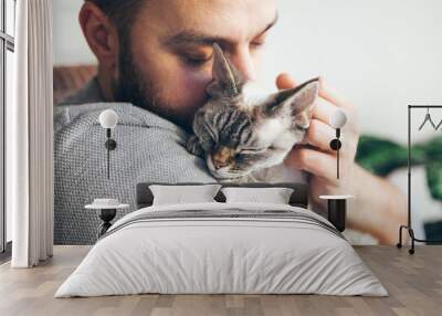 Portrait of happy cat with close eyes and young beard man snuggling. Handsome young man is hugging and cuddling his cute color point Devon Rex kitten. Domestic pets. Kitty likes attention and purrs Wall mural