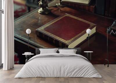 A leather bound book sits on a wooden table. The book is old and has gold embossing on the cover. The table is made of wood and has a red rug underneath it. The scene gives off a sense of nostalgia Wall mural