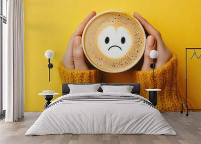 A person is holding a coffee cup with a sad face drawn on it. The cup is placed on a yellow background Wall mural