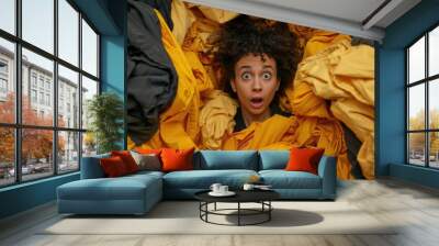 A woman is in a pile of clothes and is looking surprised. The clothes are yellow and black Wall mural